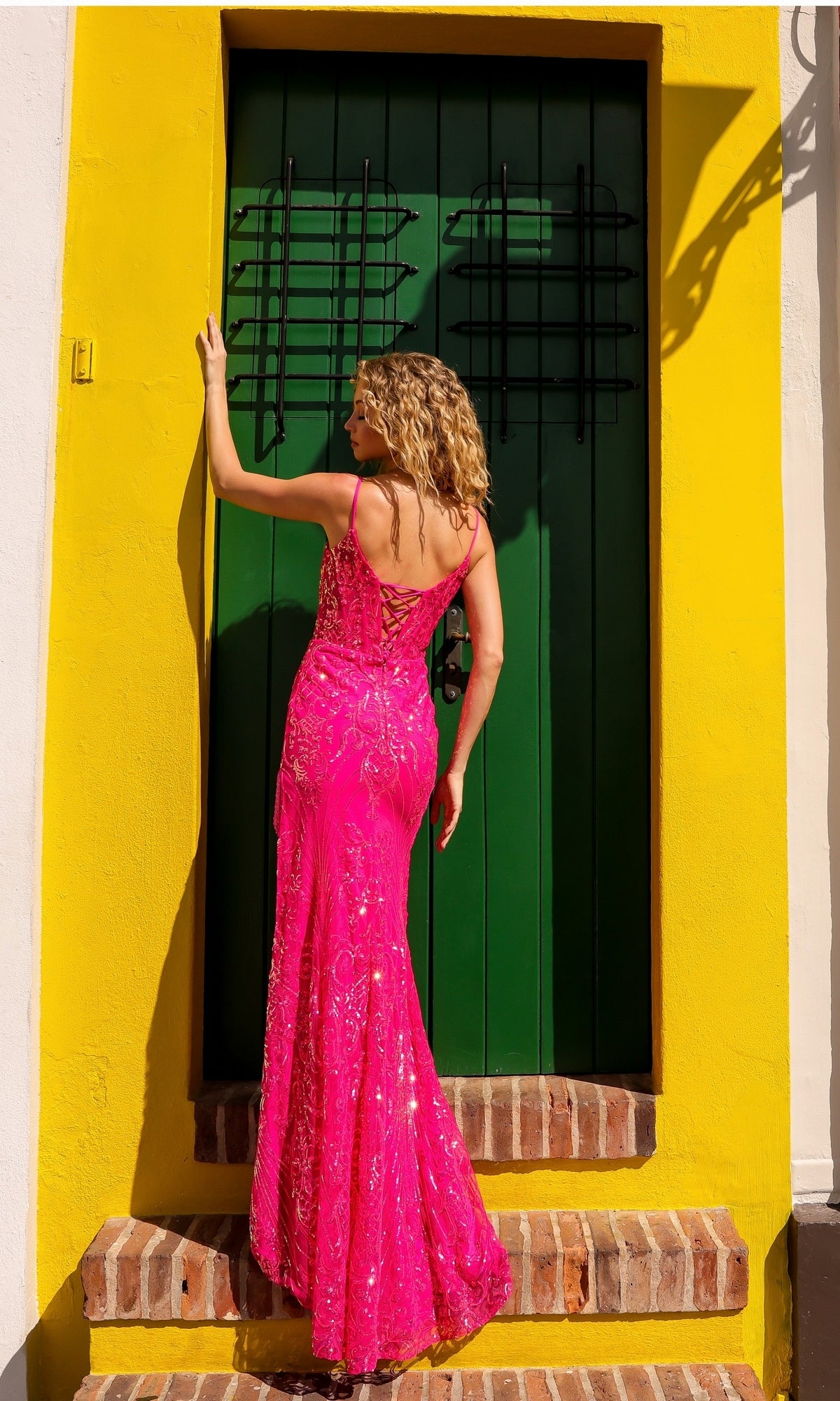 A woman wearing the Nox Anabel D1465 Fringed Long Sequin Prom Dress stands on steps facing a closed green door framed by a yellow wall.